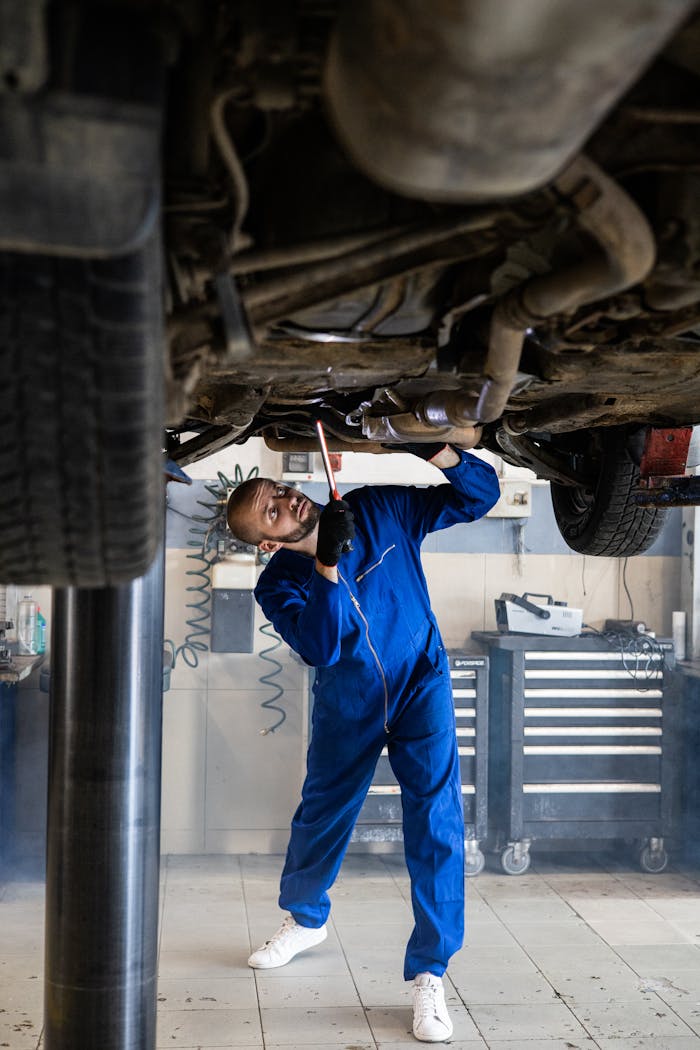 About A female mechanic in blue coveralls inspects a car's undercarriage in an auto repair shop.